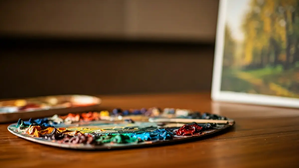 Imagen genérica de una paleta de pintor con óleos y pinceles, y un lienzo con un paisaje natural borroso.