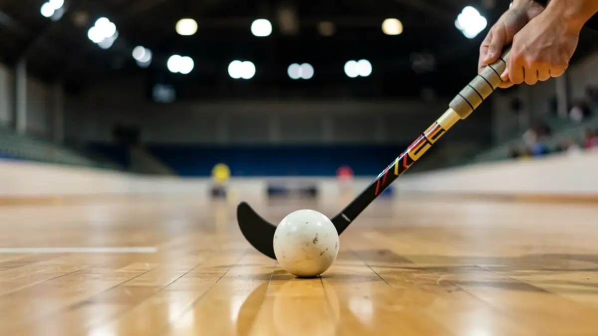 Primer plano de un stick y una pelota de floorball sobre una cancha de madera con luces de estadio desenfocadas al fondo.