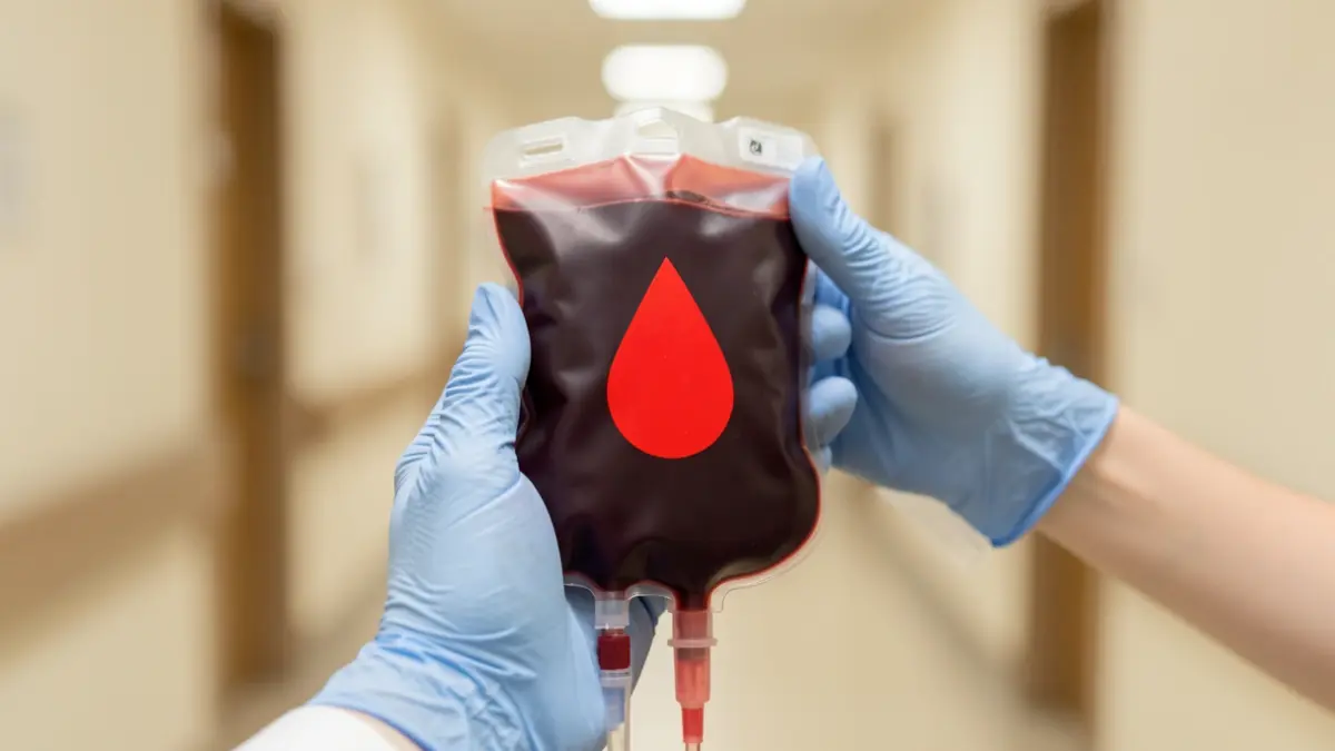 Blood bag held by gloved hands in a hospital setting.